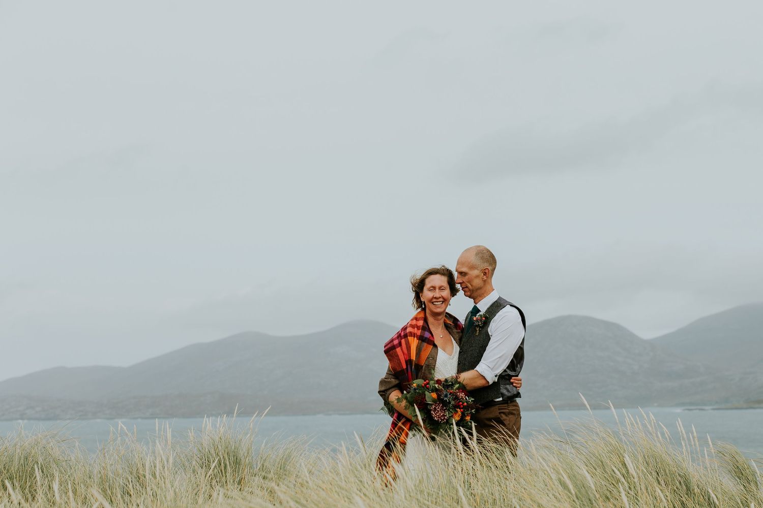 Couple embracing on grassy dunes with mountainous coastal landscape in background.