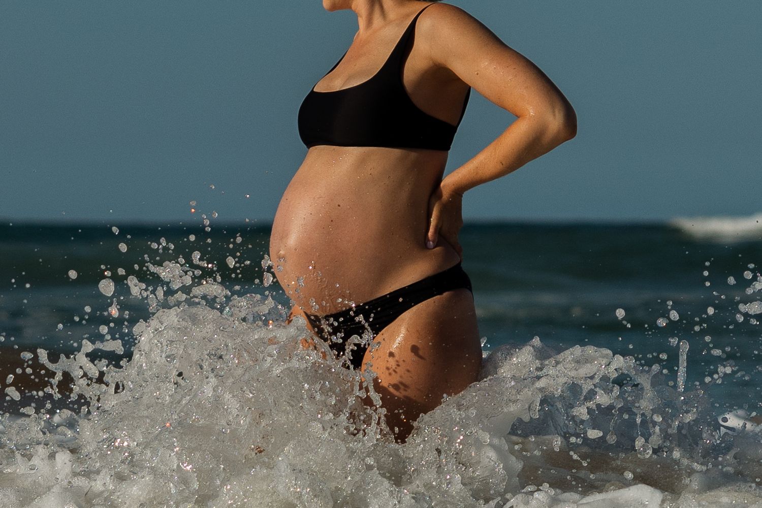 Pregnant woman in black swimwear splashing in ocean waves.