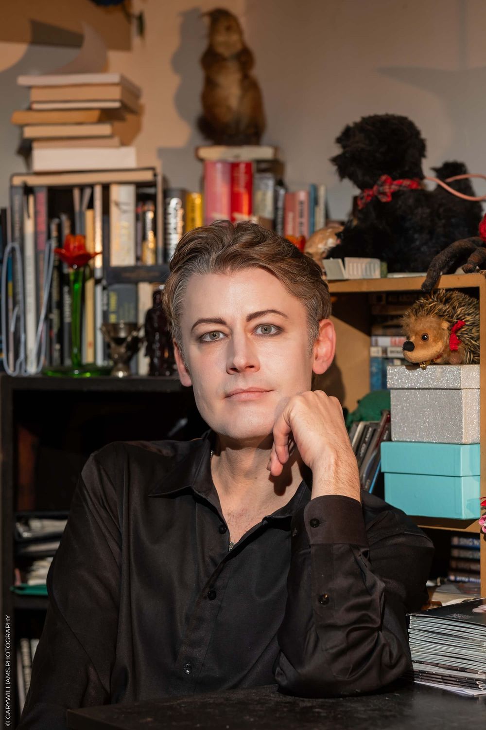 Portrait of someone seated at table wearing black shirt with bookshelves and decorative items in background.
