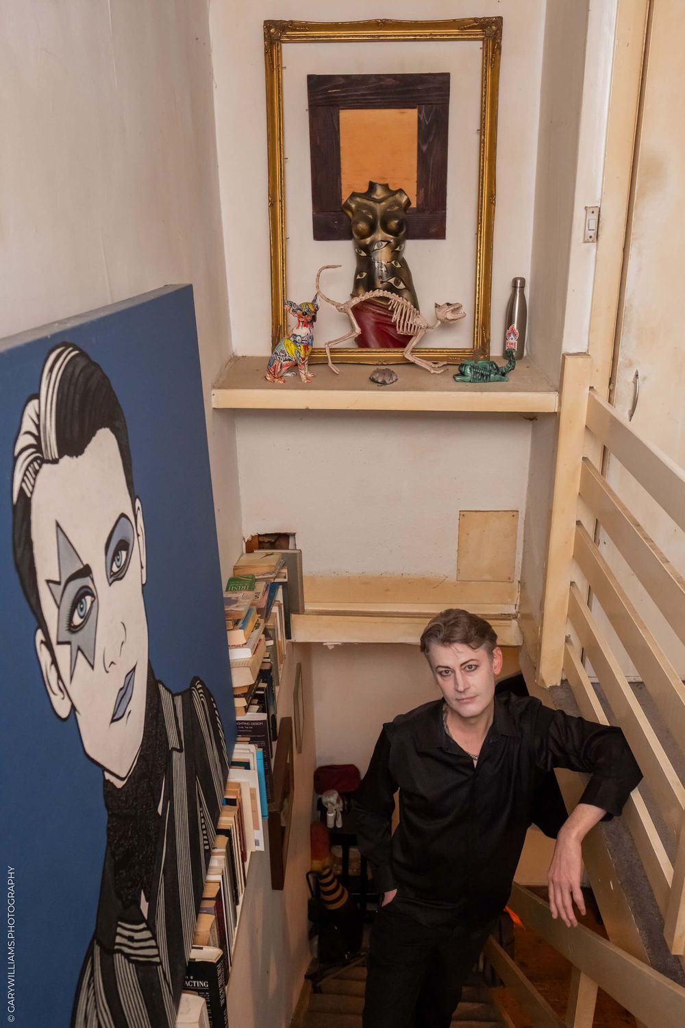 Interior stairwell with a large blue portrait on wall, wooden railing, and decorative shelf with framed artwork above.