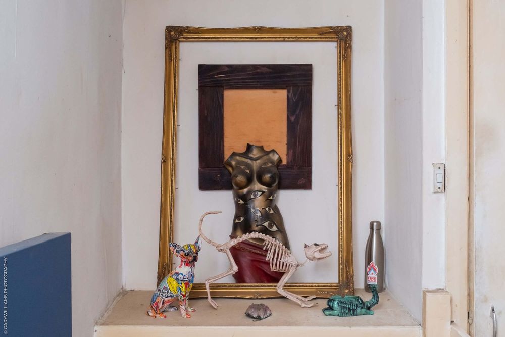 A framed artwork and decorative shrine display with colorful figurines and bottles on a white shelf.