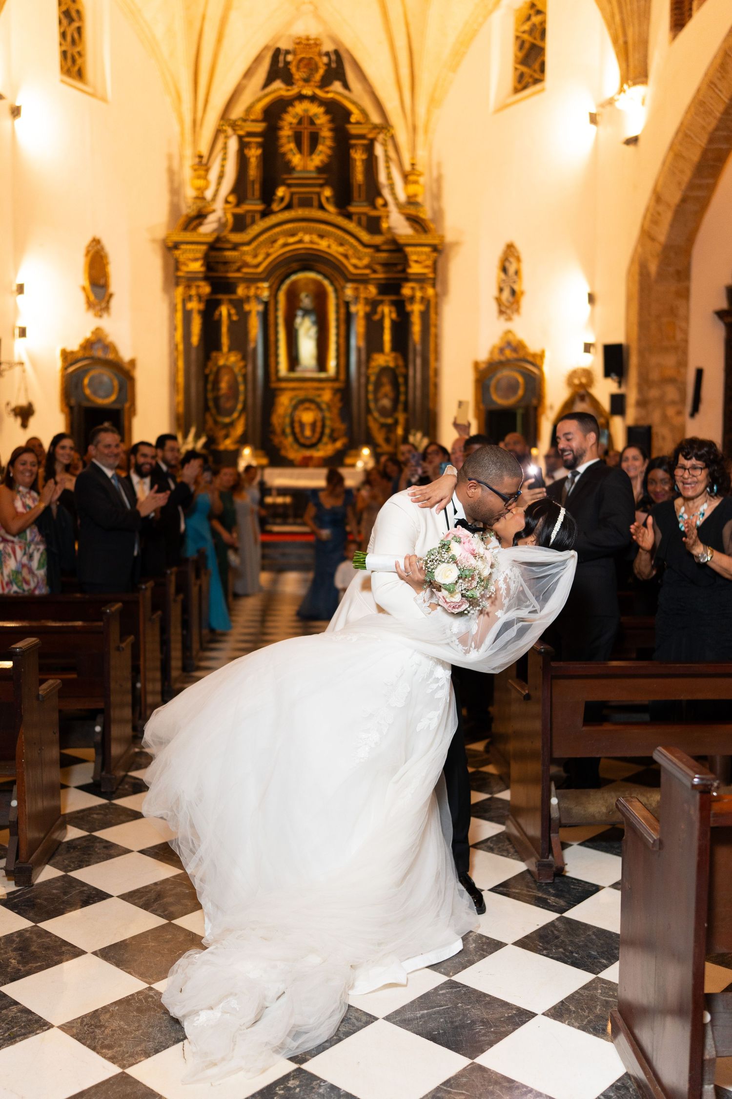 Romantic wedding dip kiss in ornate church with checkerboard floor and guests watching.
