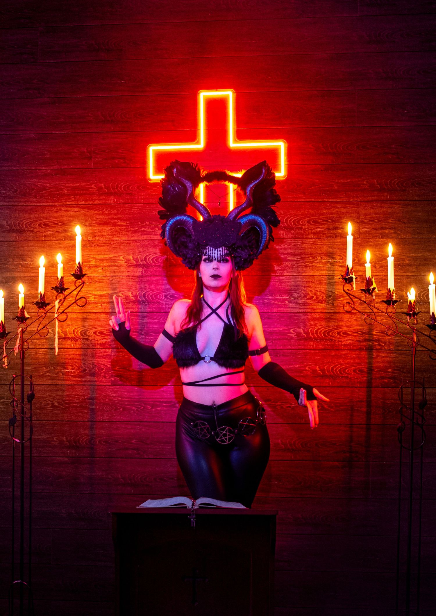 A gothic cosplay scene with candlelight and neon red cross, featuring dramatic dark attire and horned accessories.