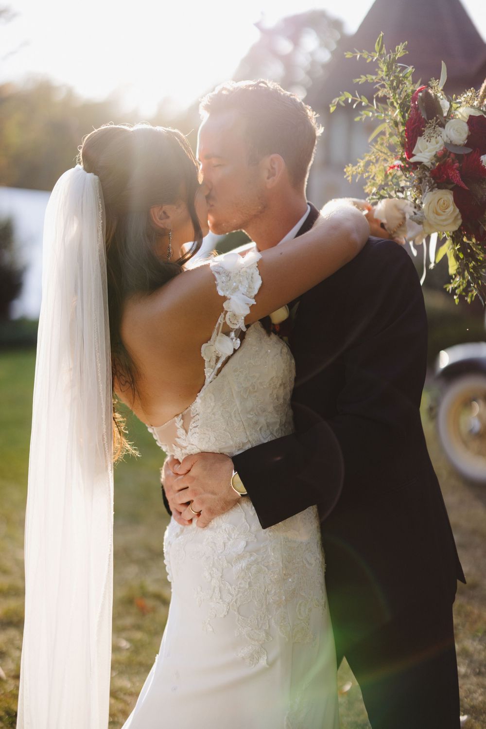 A romantic wedding couple embraces during sunset while the bride holds a beautiful floral bouquet.