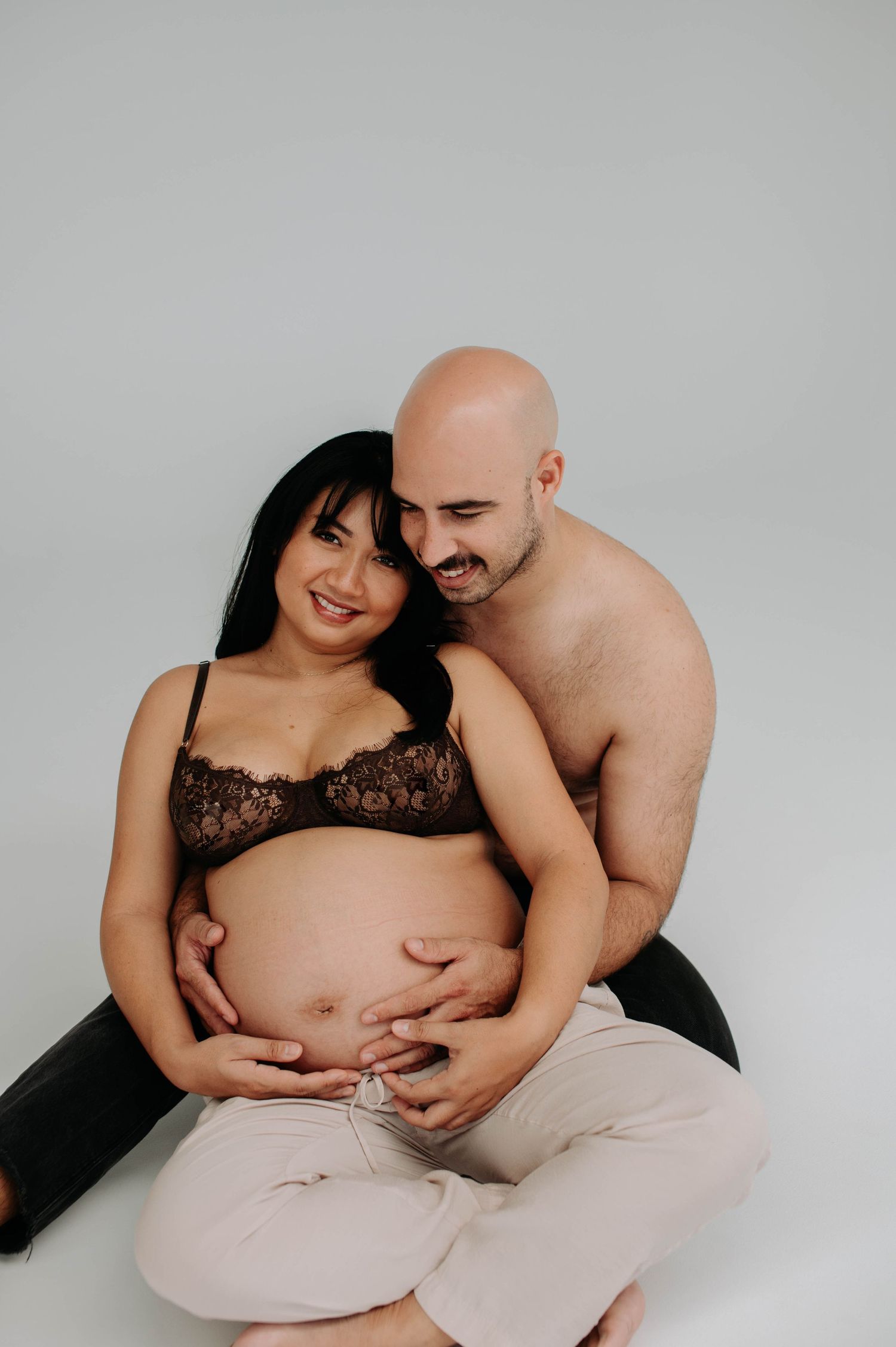 Intimate maternity photoshoot showing couple embracing on white background.