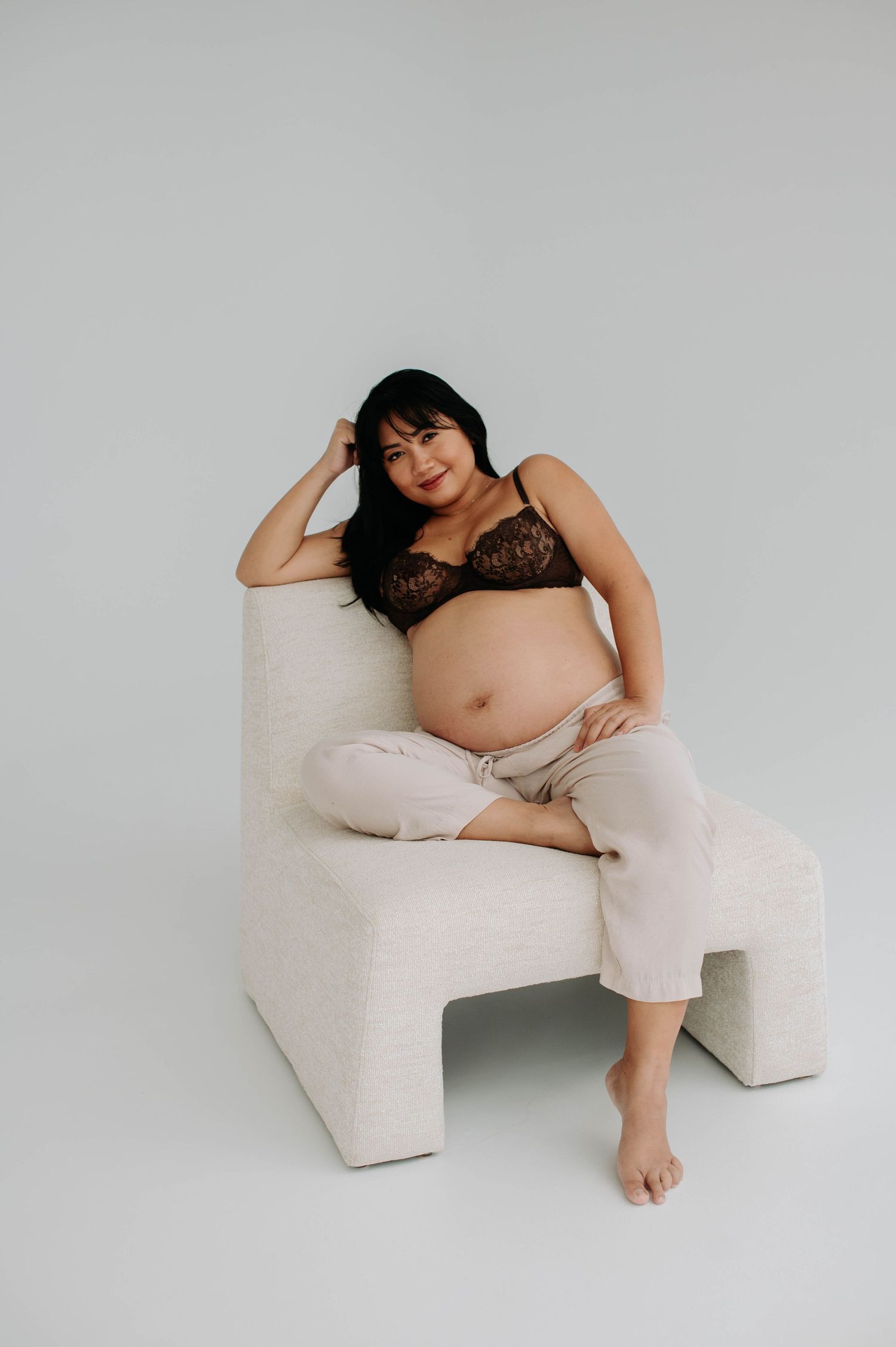 Pregnant woman in black lace bralette lounging on white chair during maternity shoot.