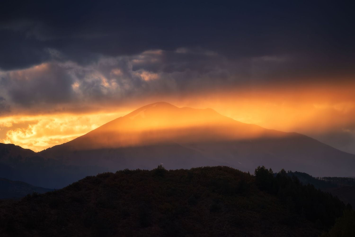 Dramatic orange sunset illuminates mountain silhouettes with dark storm clouds and golden rays of light in the sky.