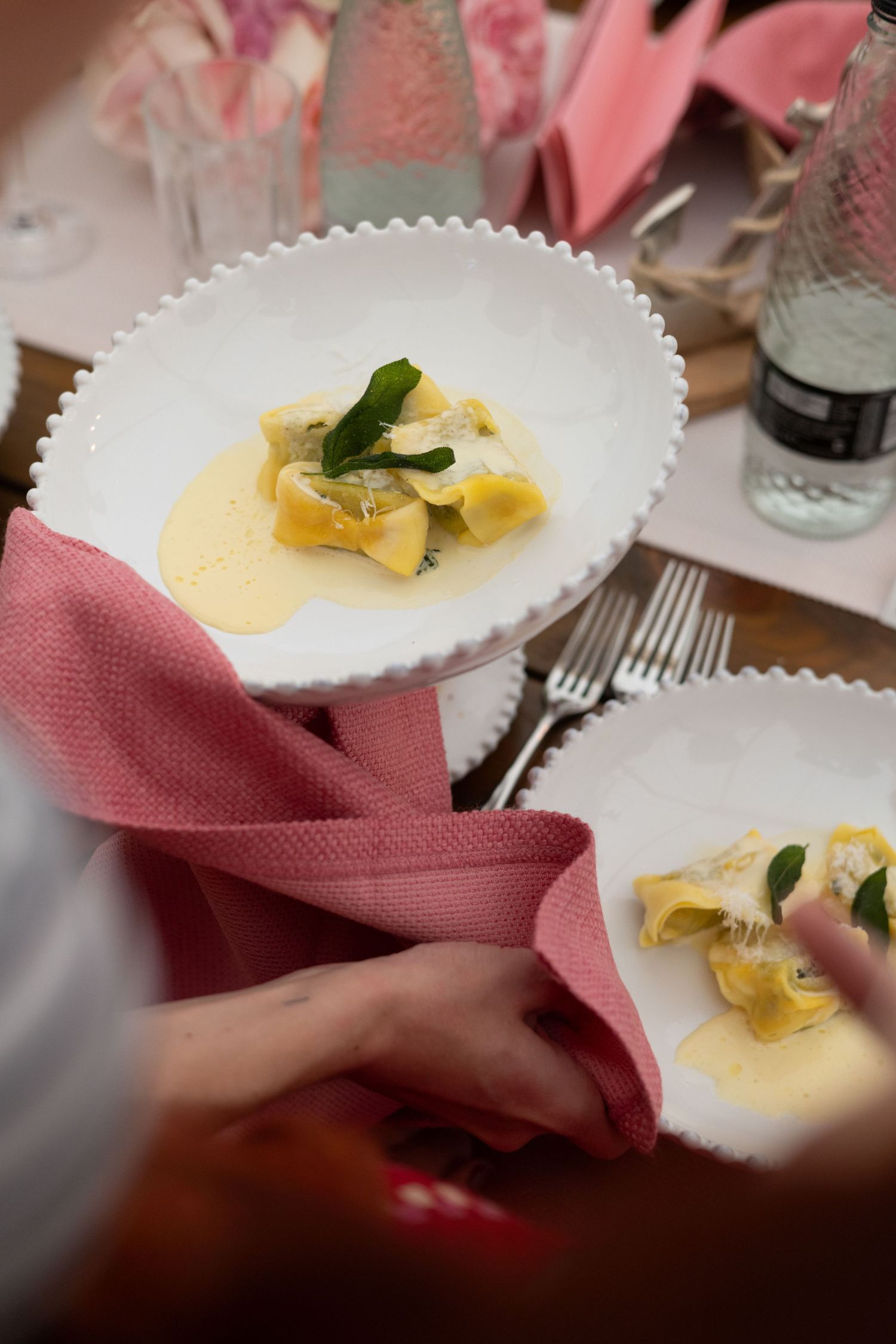 Elegant plated pasta dish with yellow sauce on white scalloped plate with pink napkin and dining setting.