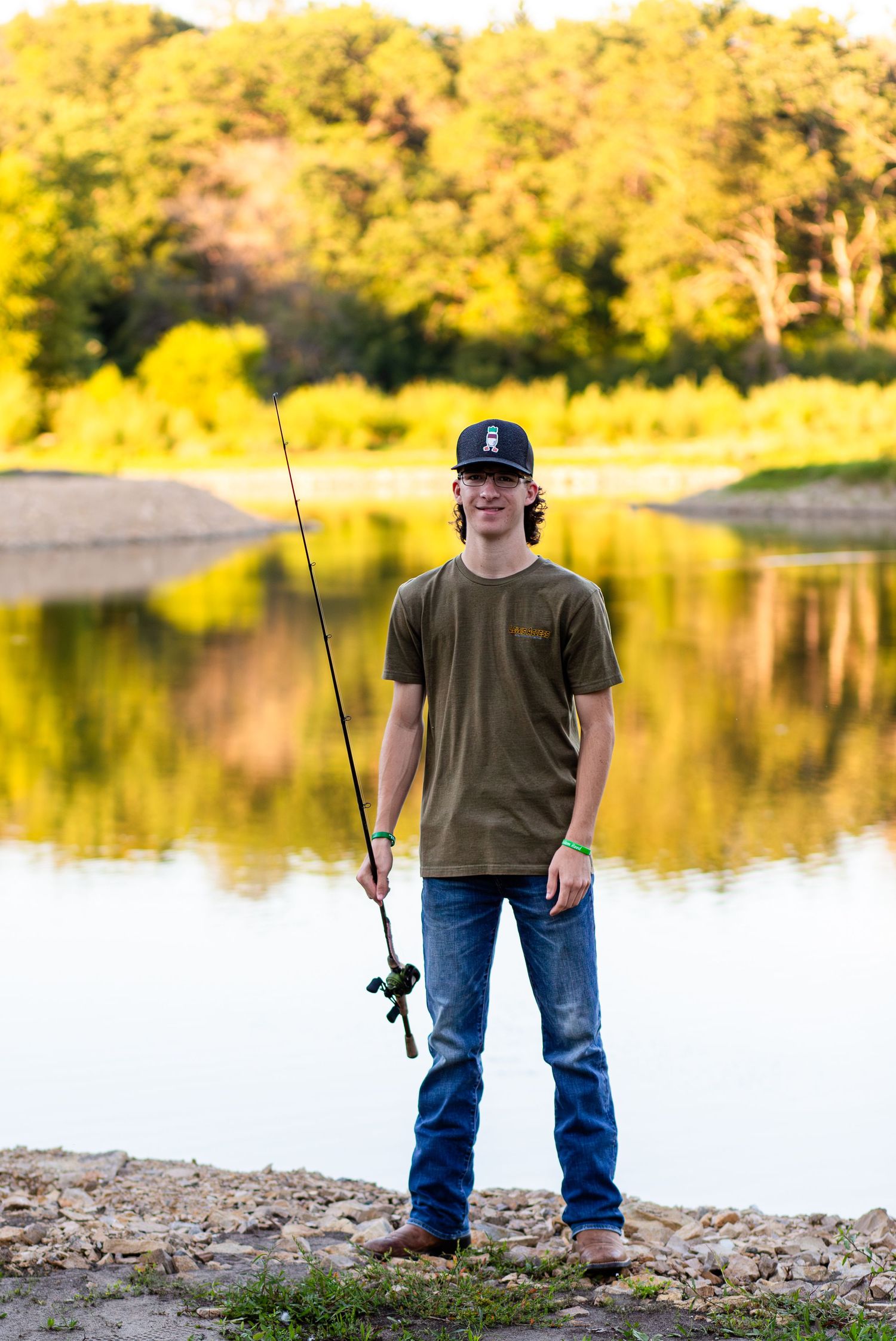 Young person in a baseball cap and casual clothes stands on a lakeshore with a fishing rod during golden sunset.
