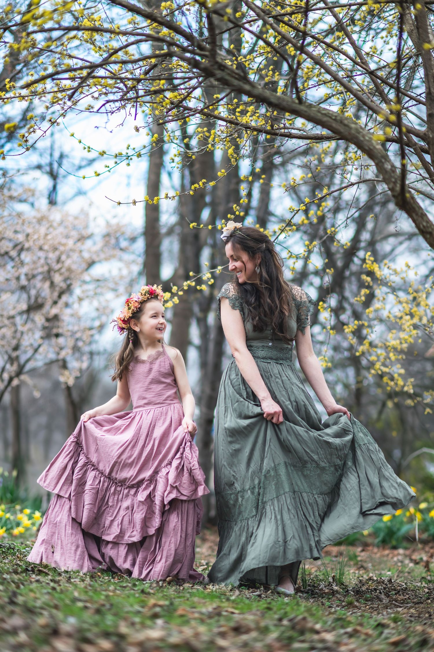Two people in vintage-style dresses twirl beneath flowering spring trees in an ethereal woodland setting.