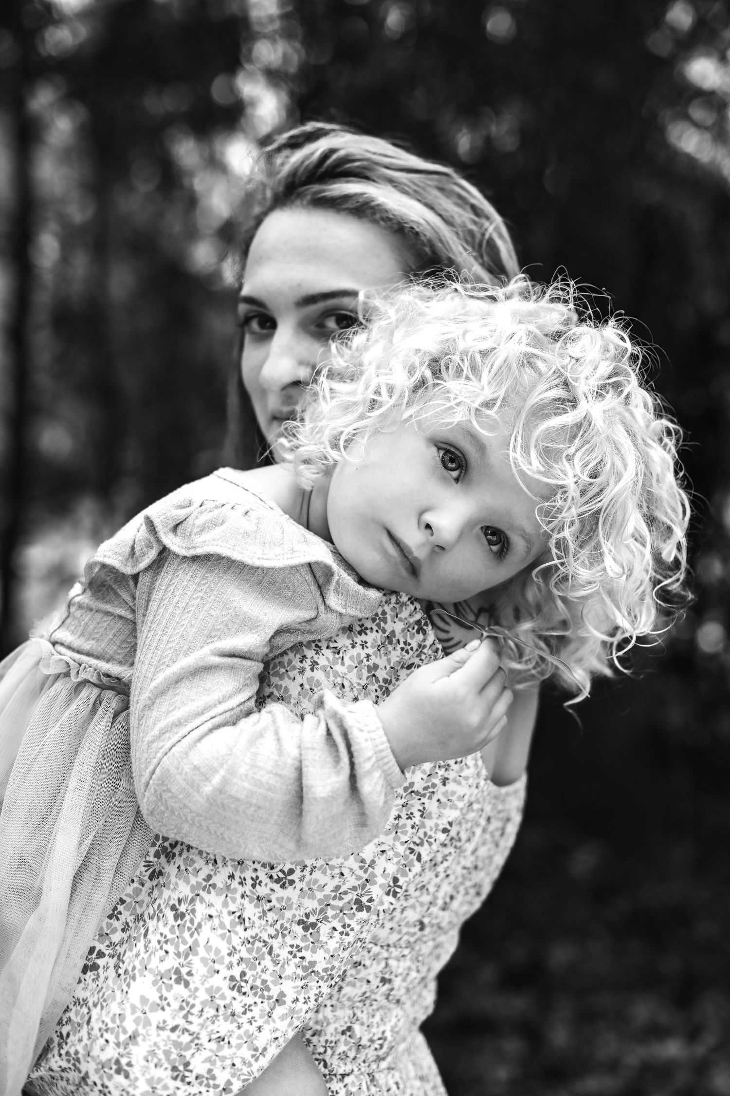 Black and white portrait showing tender embrace between parent and child with curly blonde hair outdoors.