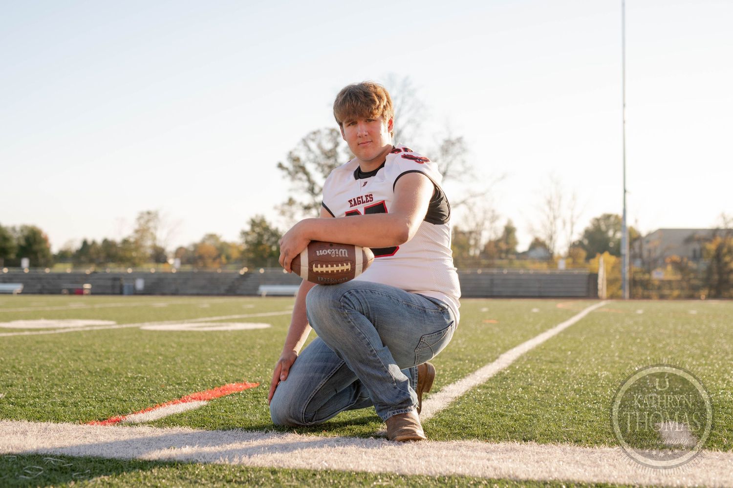 Player crouches on football field sideline holding ball during golden hour lighting.