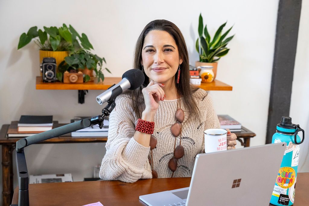 Content creator recording a podcast at home desk setup with microphone, laptop and plants on wall-mounted shelves.