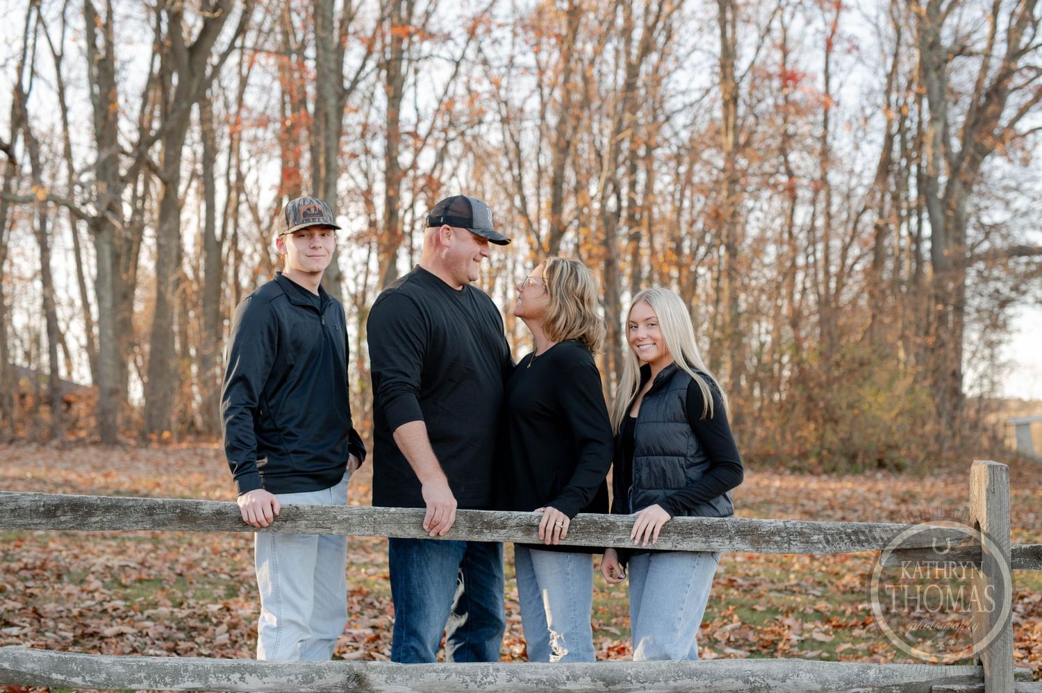 Small group poses casually along wooden fence rail in autumn woodland setting.