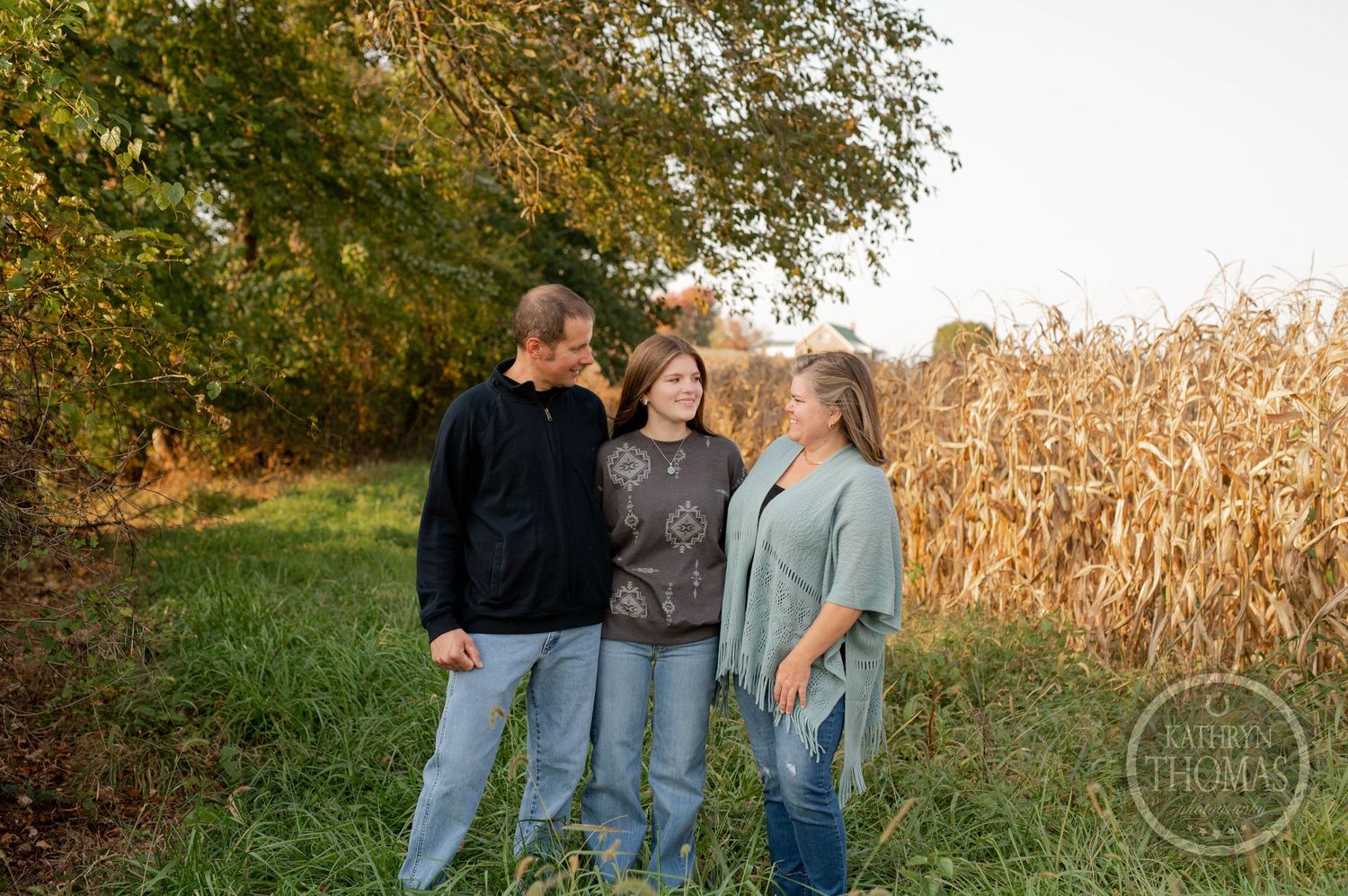 Family of three smiling together in front of autumn cornfield.