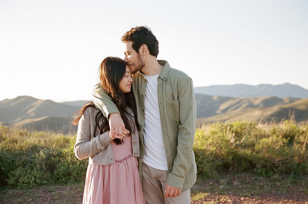 A couple shares an intimate moment while standing together in a grassy field with mountains in the background at sunset.