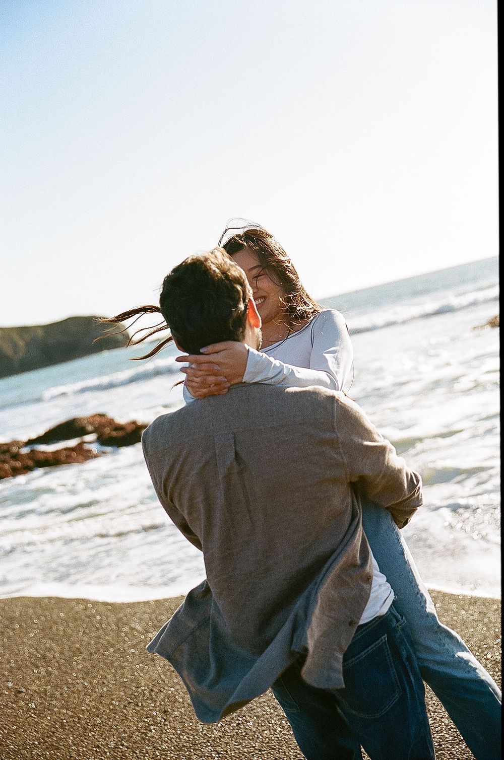 A romantic couple embrace and kiss on a sunny beach with ocean waves crashing in the background.