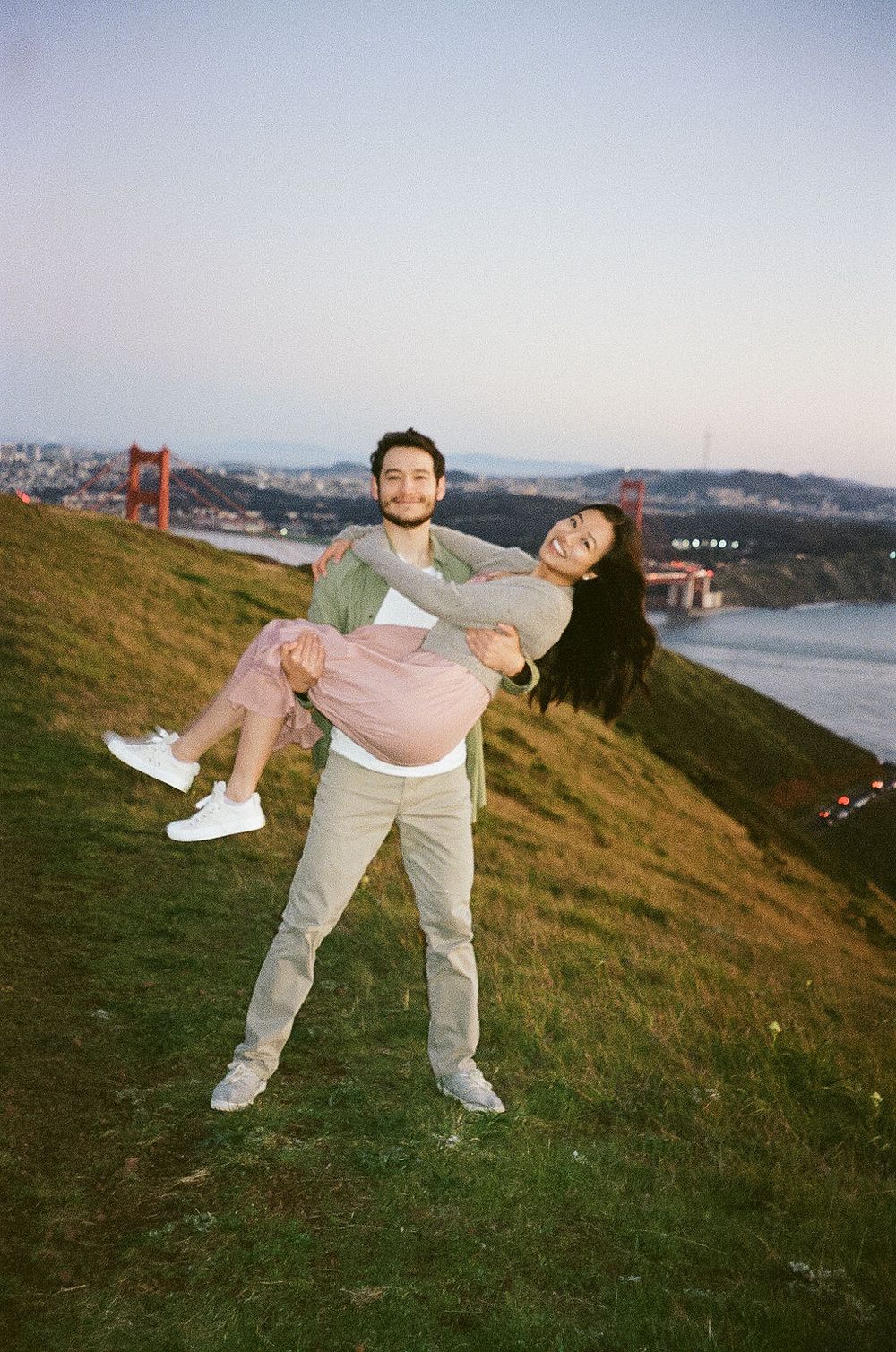 A playful couple poses on a grassy hillside overlooking the Golden Gate Bridge in San Francisco.