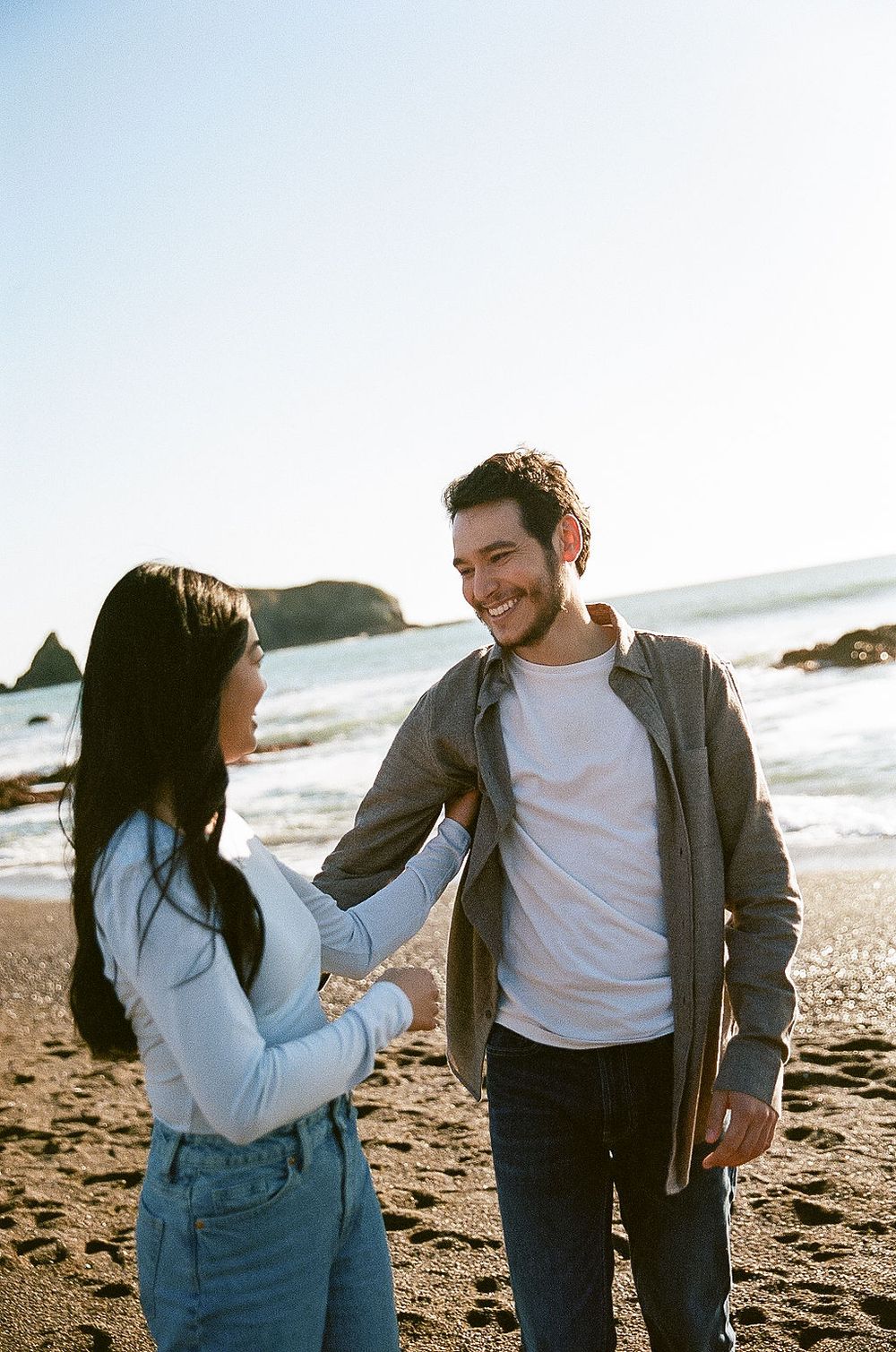 A couple shares a joyful moment together while standing on a beach with coastal rock formations visible.