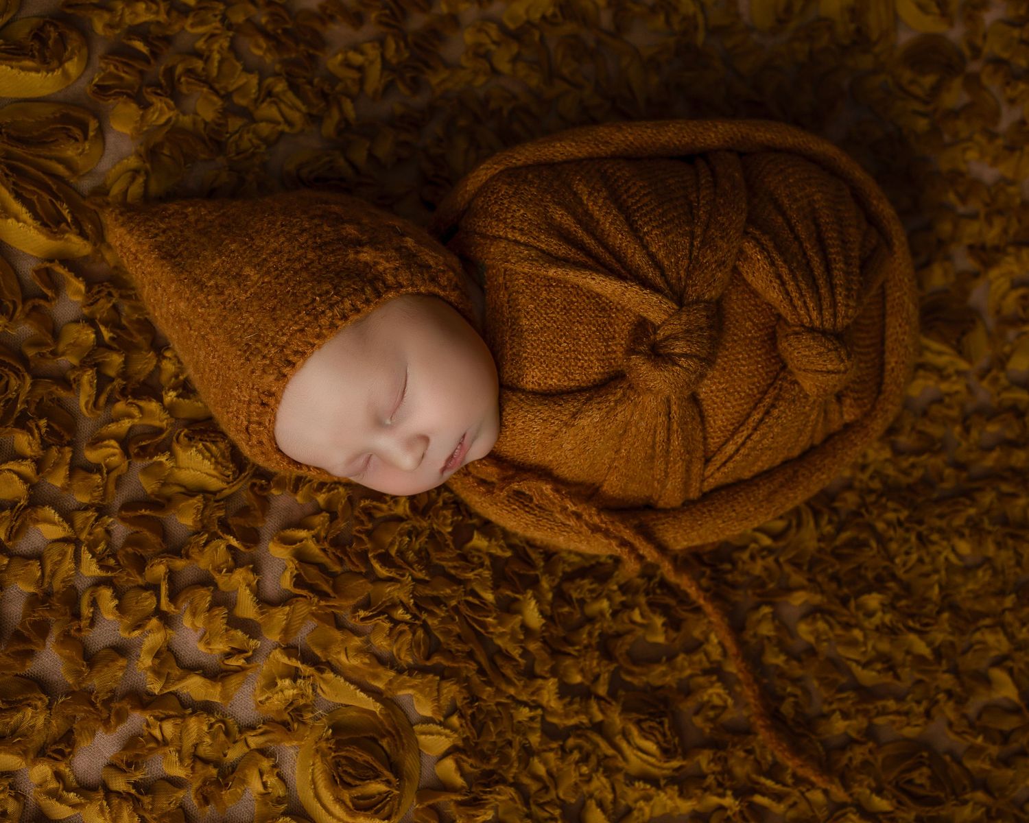 Sweet newborn baby wrapped in golden knit swaddle sleeping peacefully on patterned golden fabric.