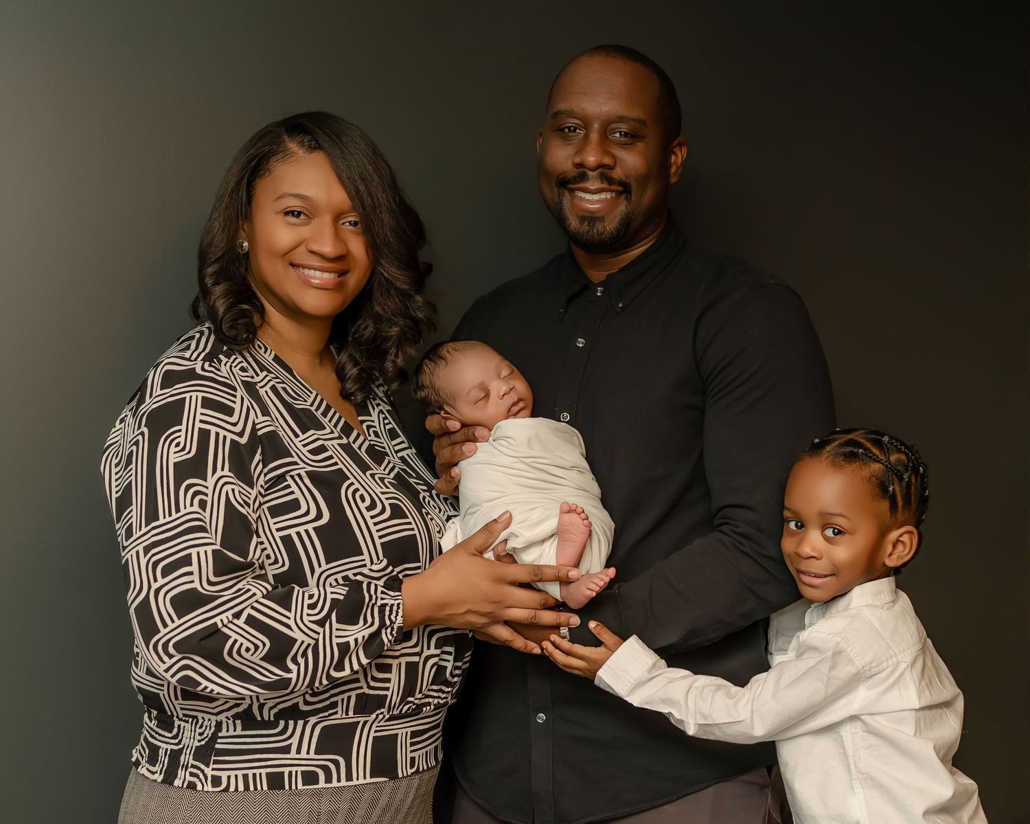 Professional family portrait with geometric patterned dress and newborn baby against dark background.