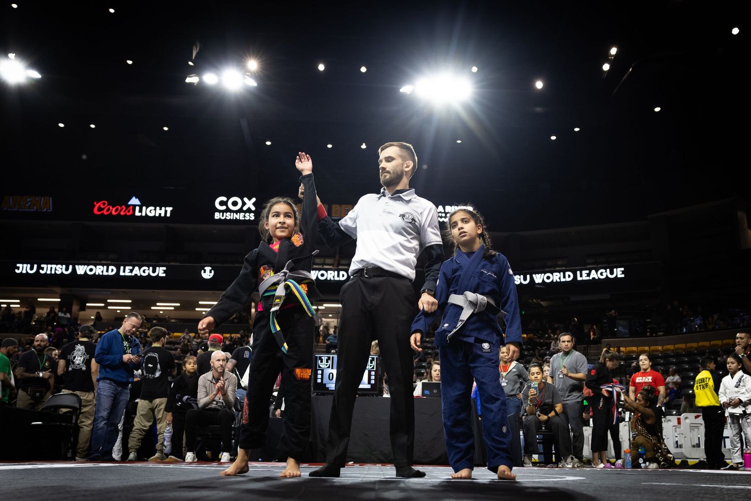 Athletes competing in a Brazilian Jiu-Jitsu tournament under bright arena lights. Winner has hand raised