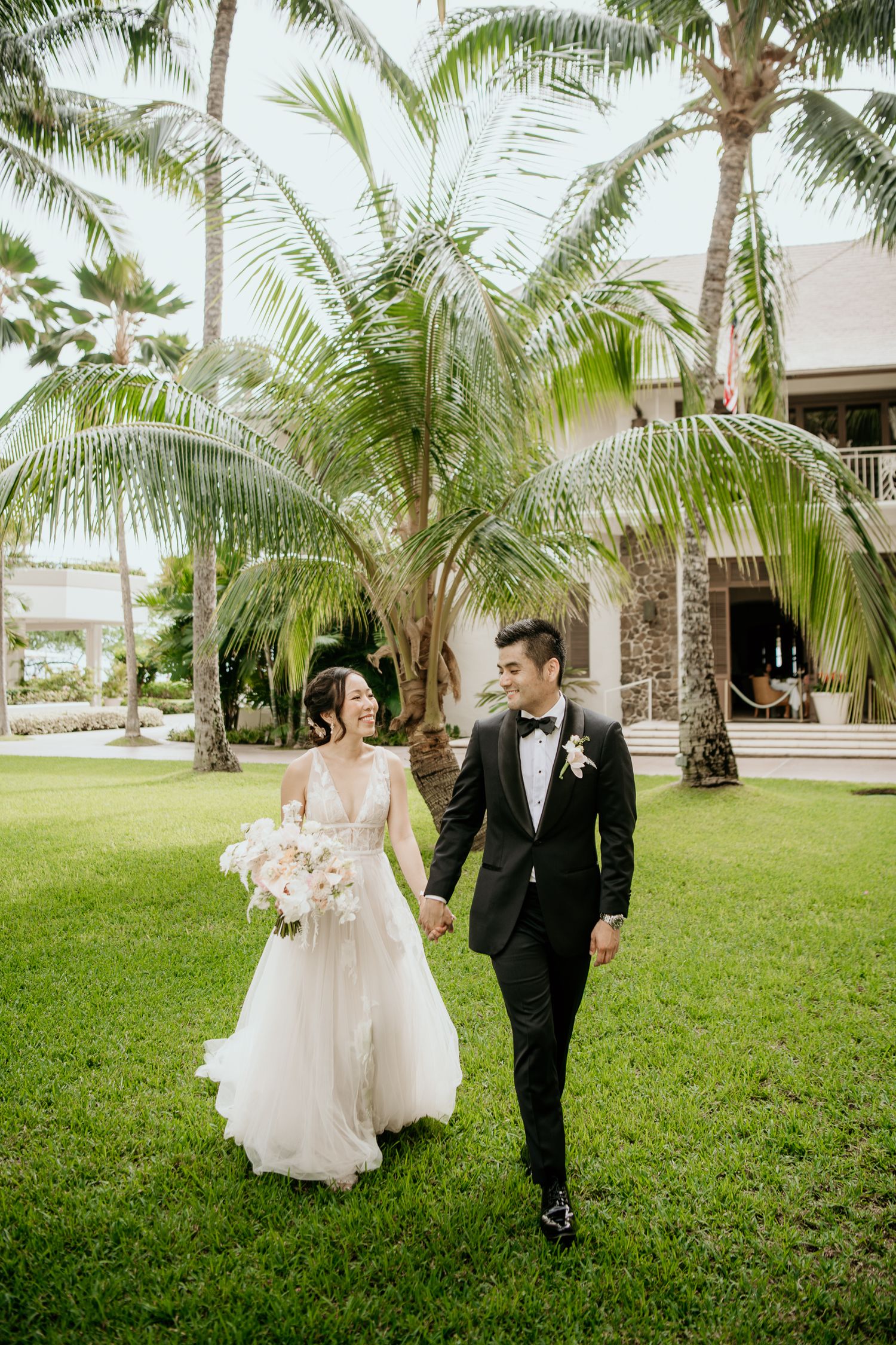 A couple walks together on a manicured lawn with tropical palm trees and a building in the background.
