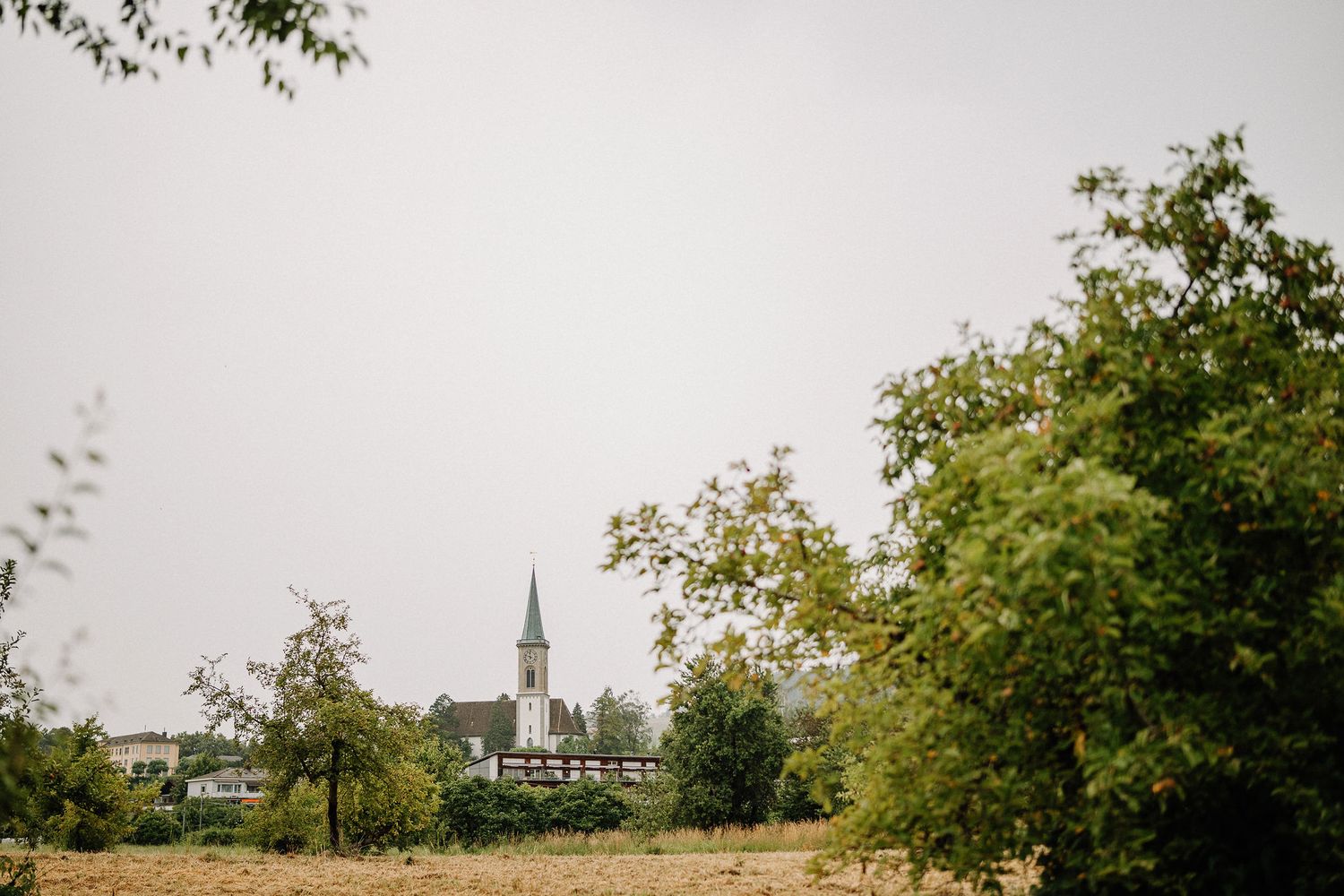 A church steeple rises above trees in a rural landscape under an overcast sky.