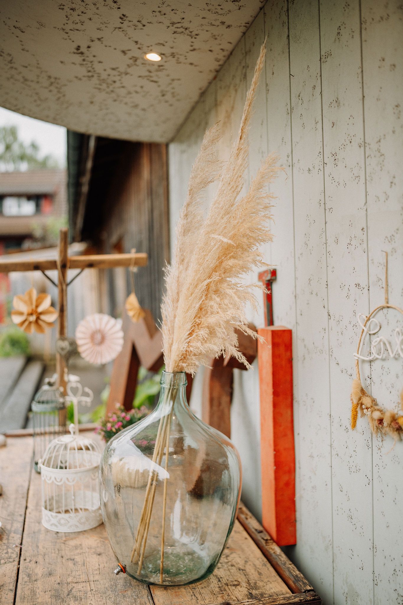 A rustic porch display with pampas grass in a glass bottle, wooden decor and candleholders against a weathered wall.