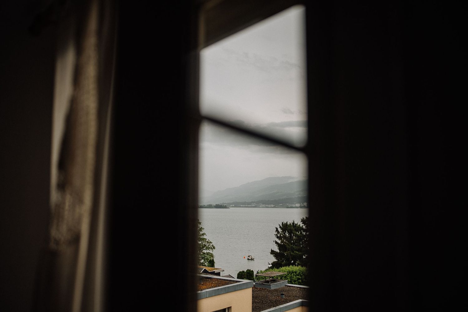 Moody lake view through a dark window frame with mountains in the distance and trees along the shore.