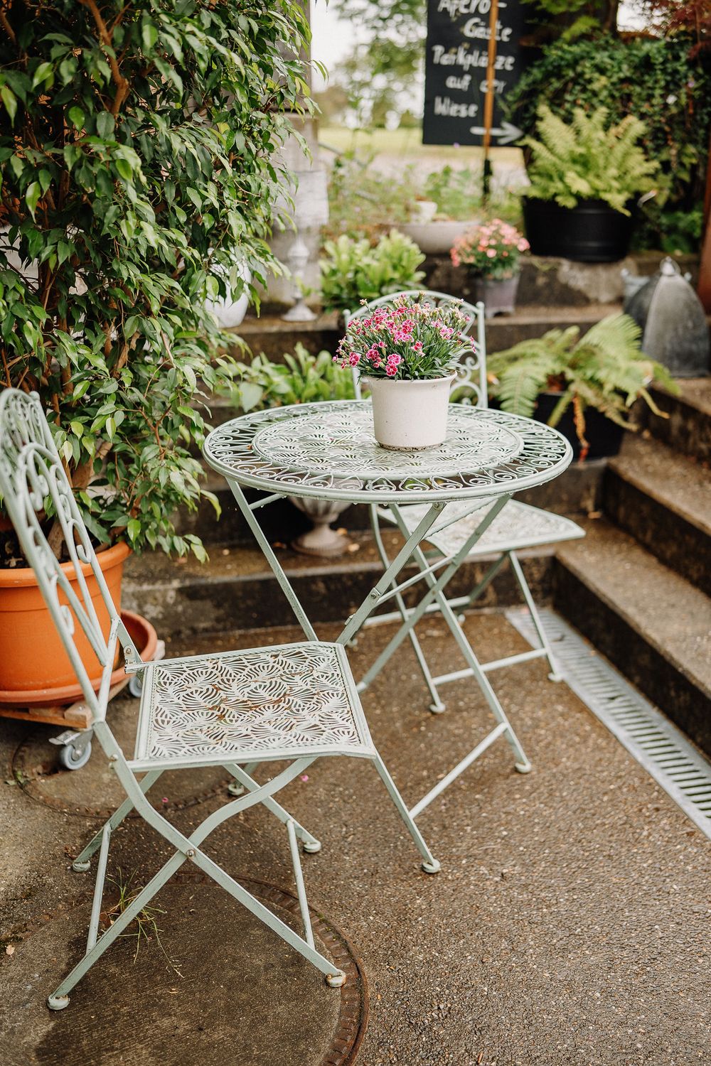 Vintage white metal bistro table and chair set with pink flowers in pot on garden patio with lush greenery background.
