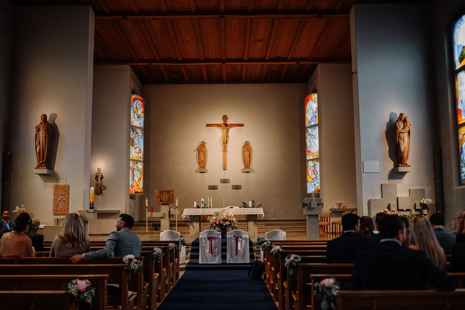 Interior view of a Catholic church with wooden pews, stained glass windows, and a crucifix on the altar during a service.