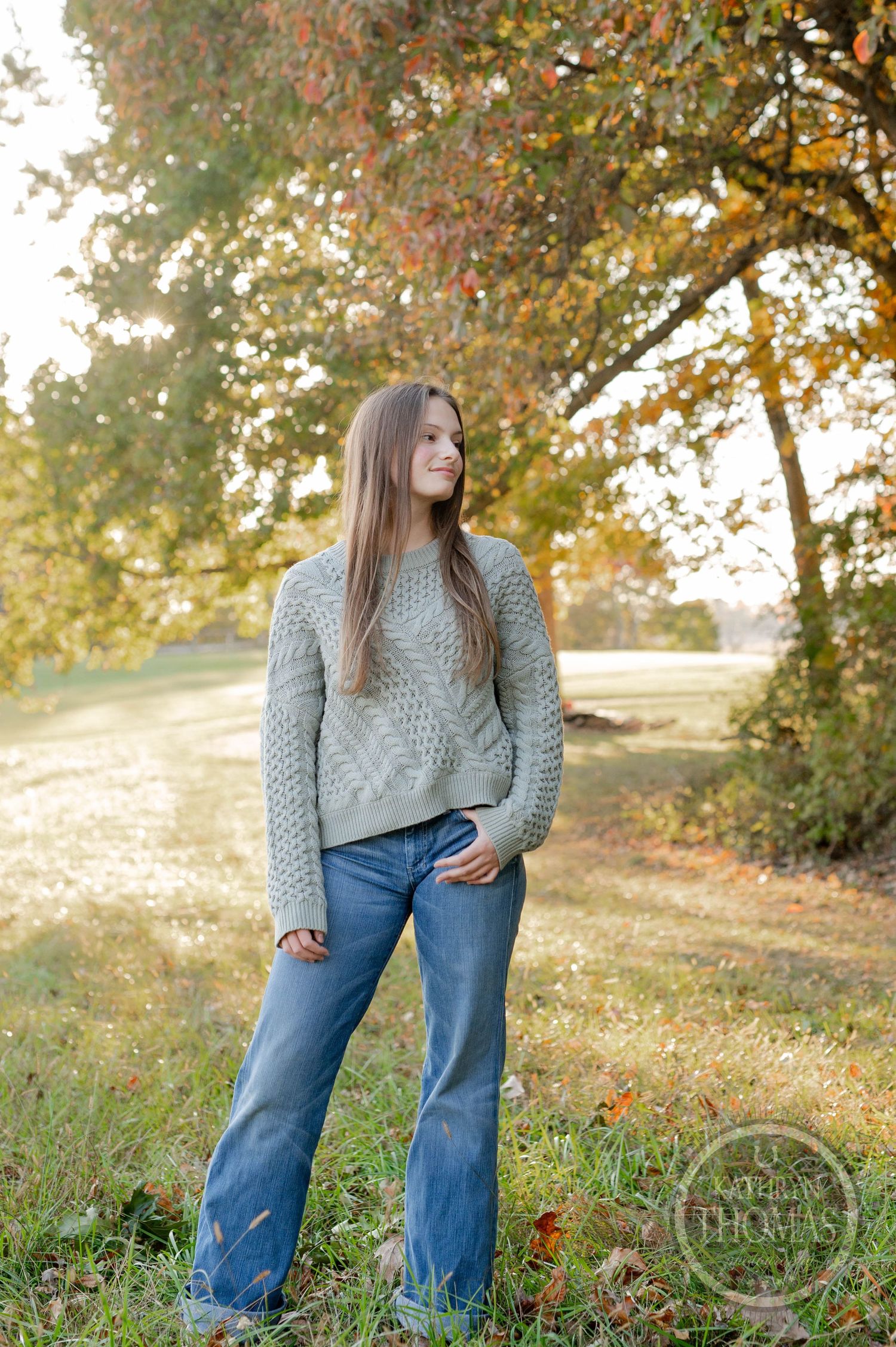 A person in a cable knit sweater and jeans poses against a golden autumn background.