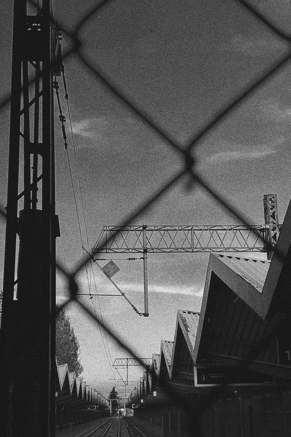 Industrial railway infrastructure viewed through chain-link fence in black and white.
