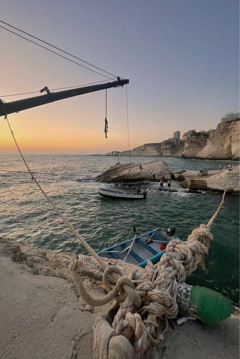 Crane and ropes at a coastal harbor during sunset with boats and limestone cliffs in the background.