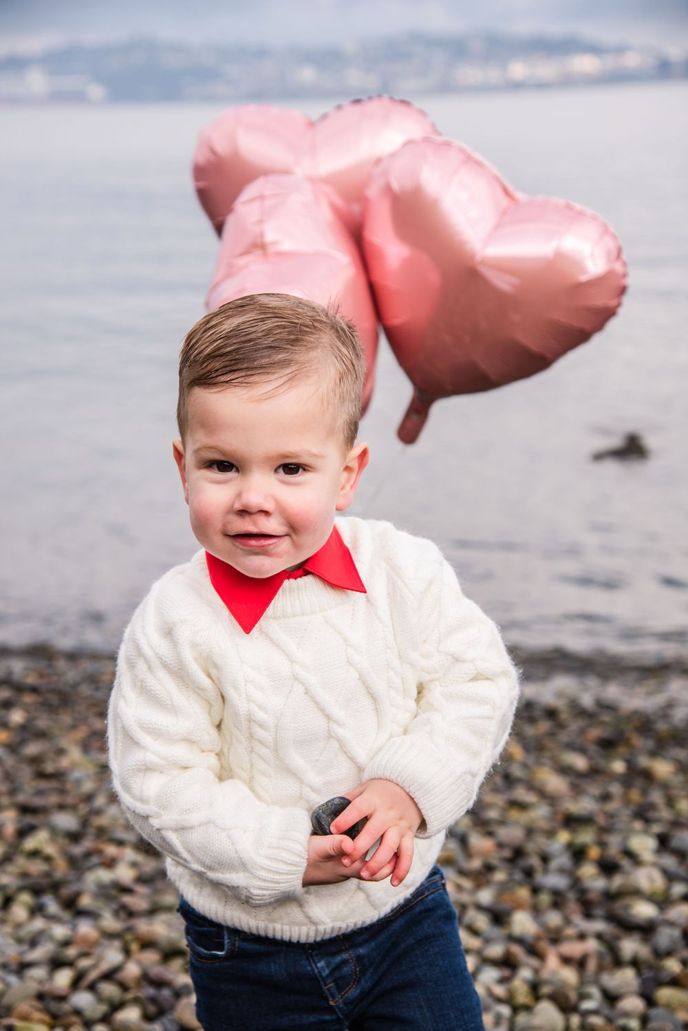 Small child in white sweater with pink balloon at rocky beach.