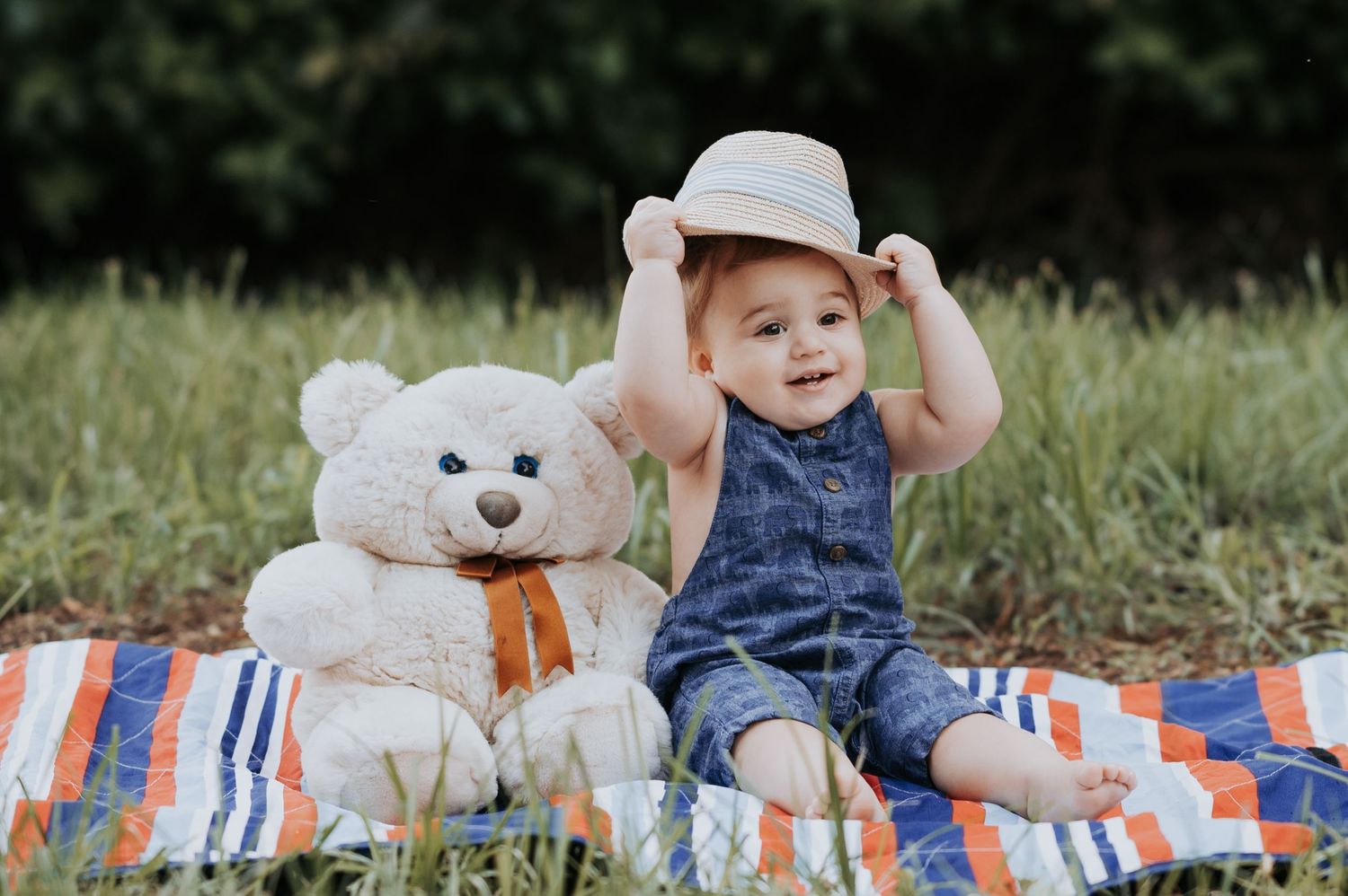A baby lies on a blanket outdoors with plush teddy bears and colorful striped pillows.