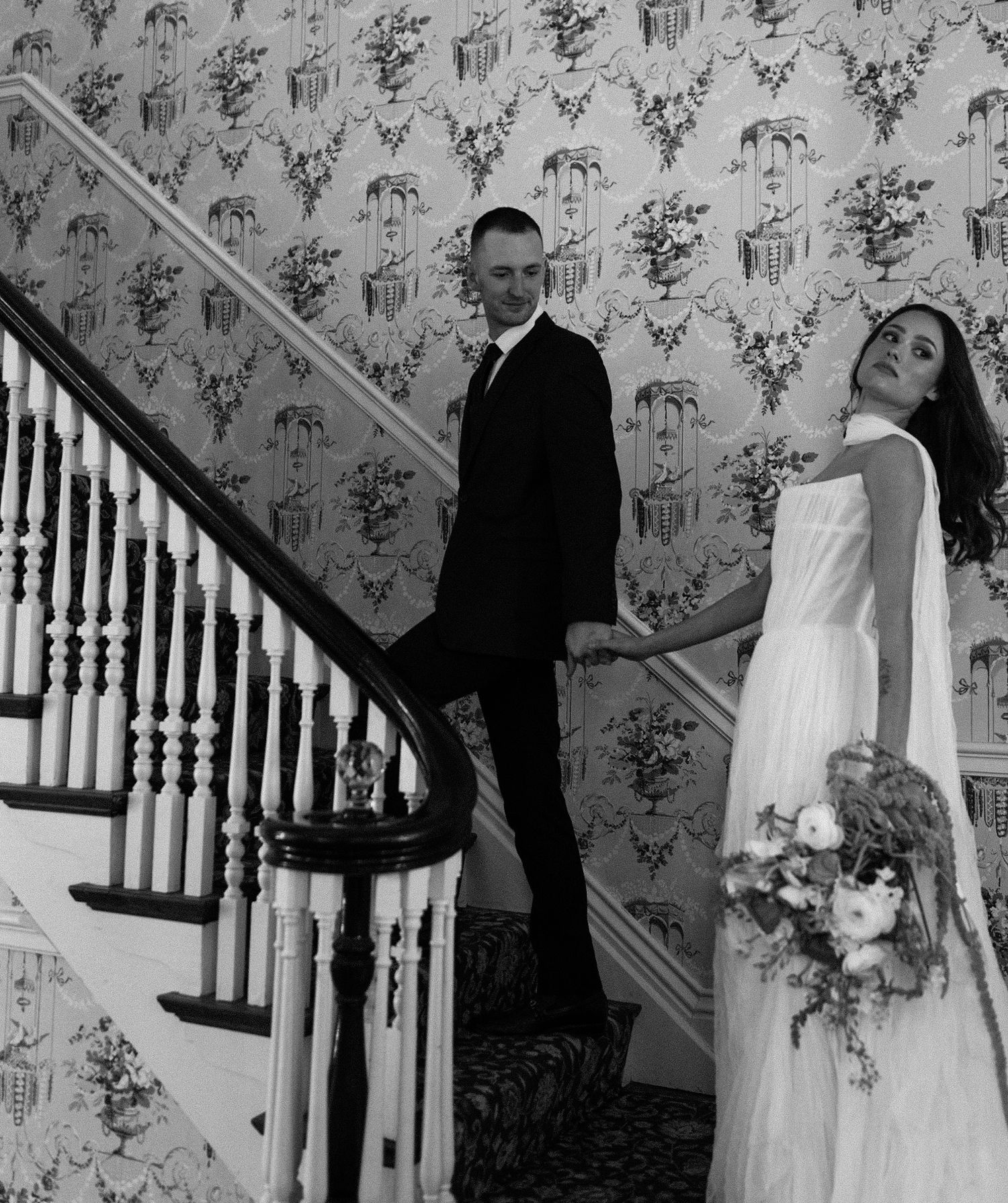 Black and white wedding photos showing elegant moments along a staircase with floral wallpaper.