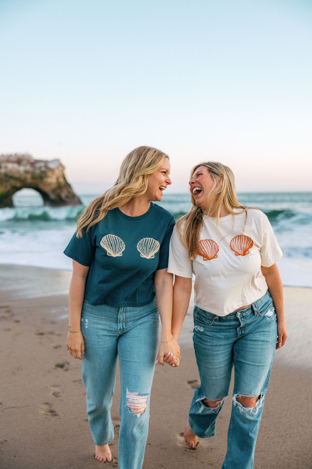 Two friends laughing together on beach wearing matching seashell t-shirts and ripped jeans at sunset.