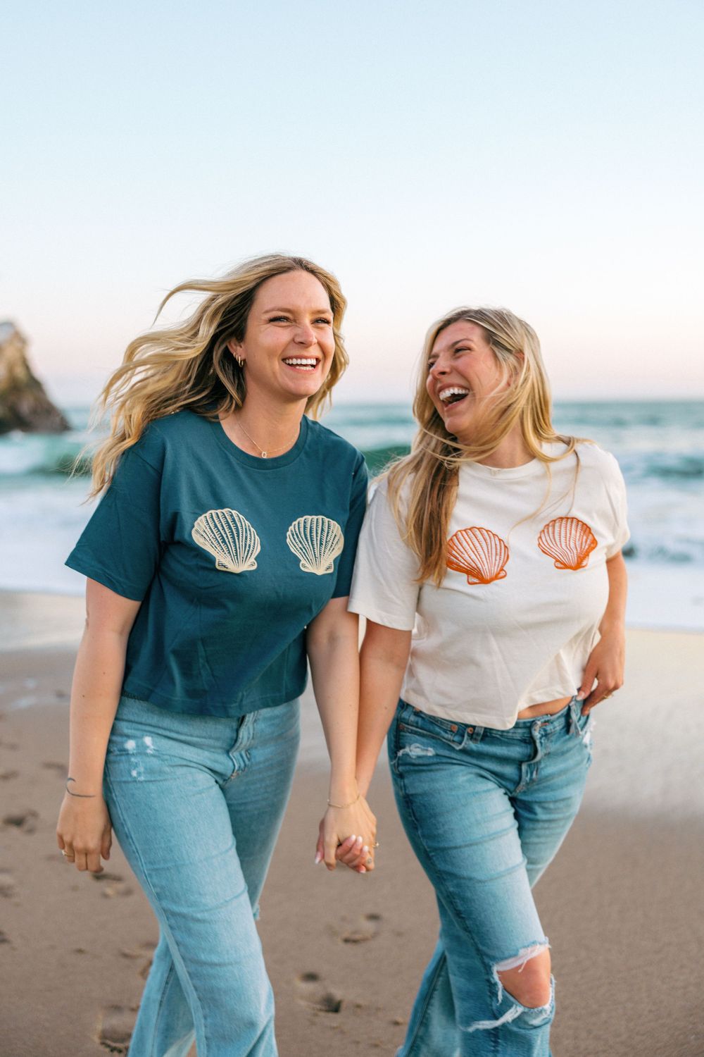 Two friends in matching seashell t-shirts laugh while walking on a beach at sunset.