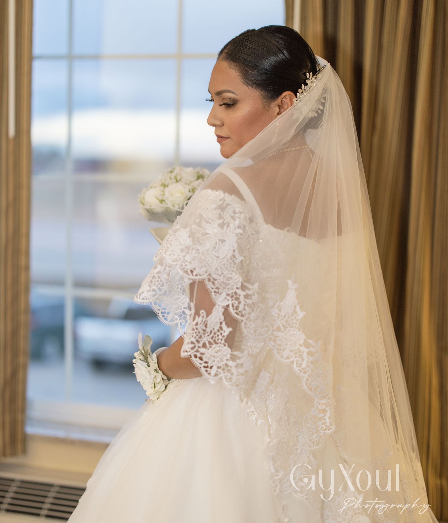 Side view of bride in lace wedding dress with veil against window light.