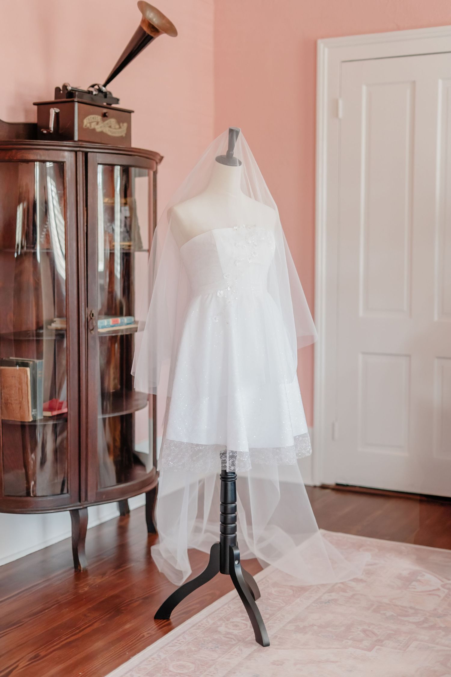 A vintage phonograph sits atop a display cabinet beside a white wedding dress on mannequin against pink walls.