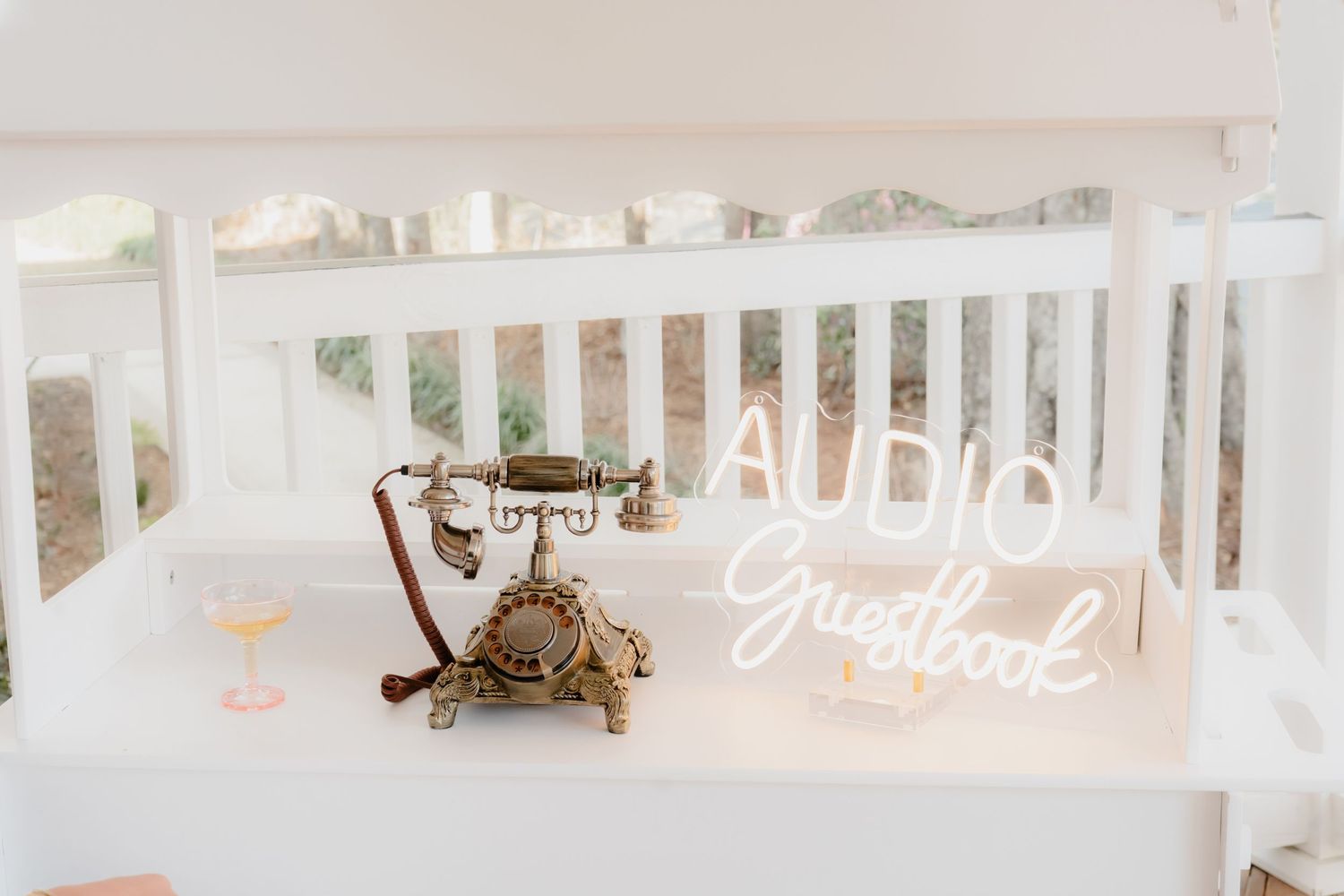 Vintage rotary telephone and glowing neon sign displayed on white porch railing with martini glass.