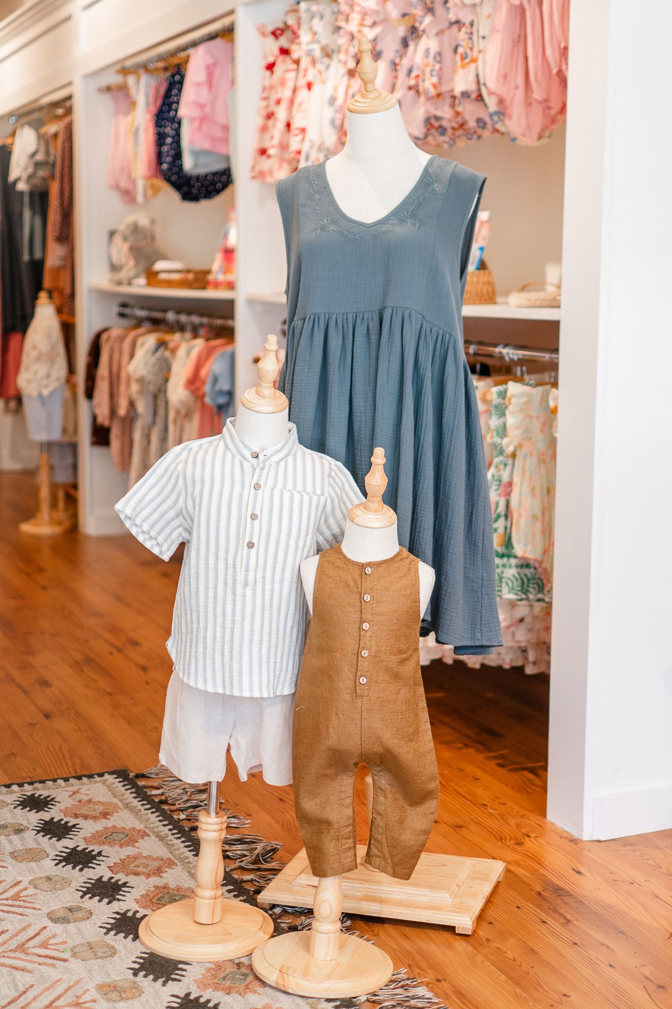 Children's clothing display with striped shirt, brown overalls and blue dress on mannequins in a boutique store interior.