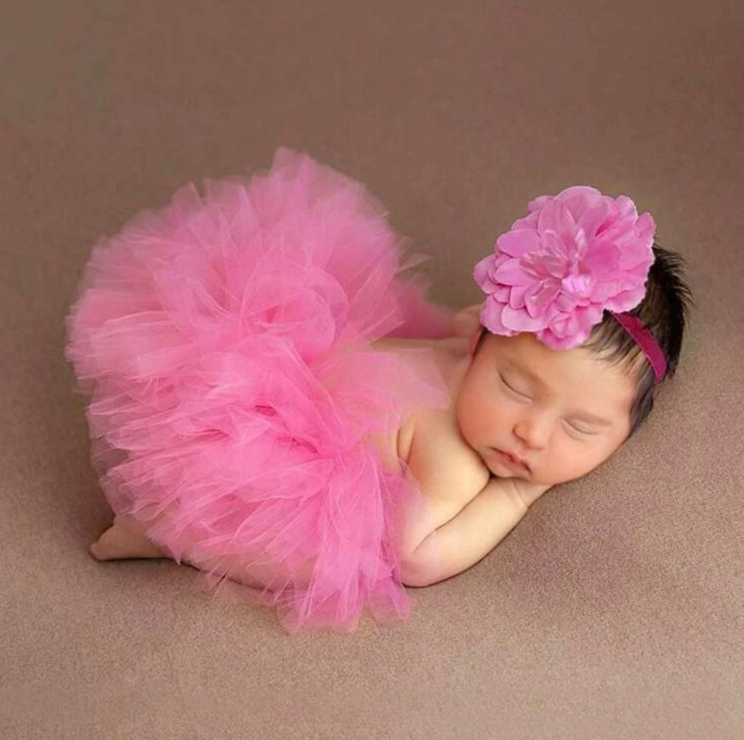 Sleeping newborn wearing pink tutu and floral headband on brown backdrop.