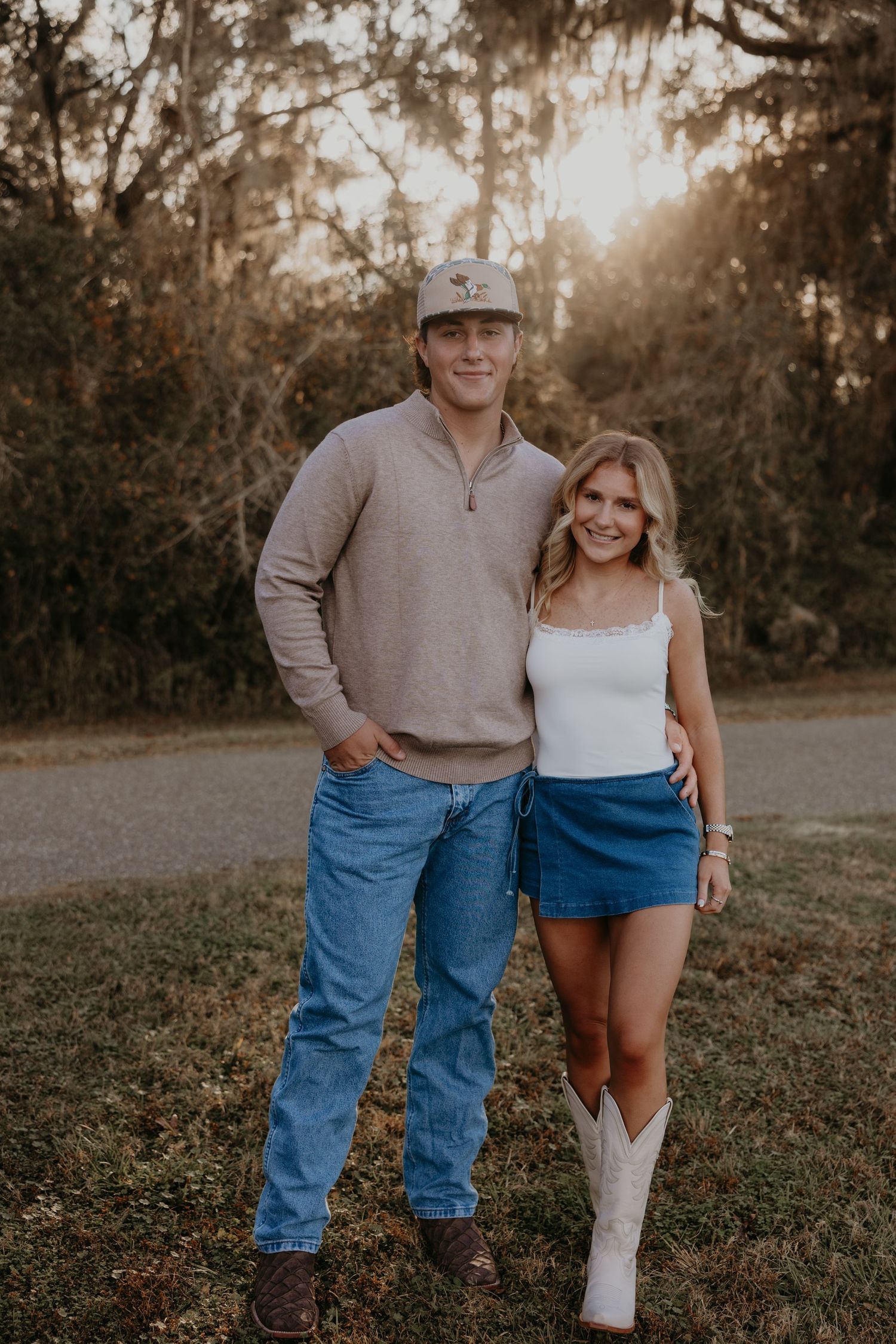 A couple poses outdoors at golden hour, man in cap and jeans, woman in denim skirt and white cowboy boots.