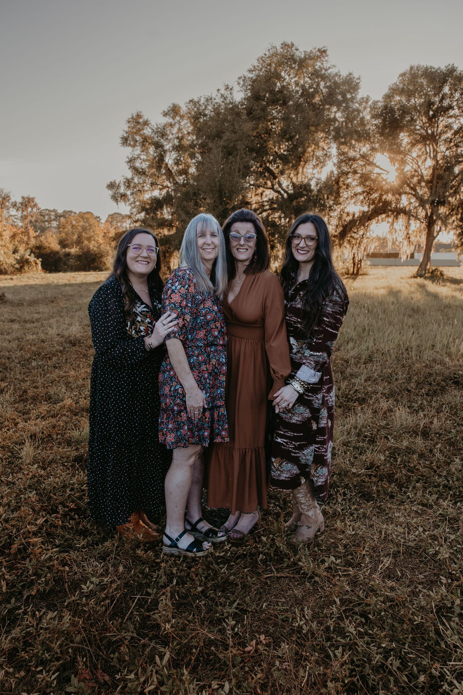 Four women posing together in a golden field at sunset, wearing stylish dresses in an outdoor fall setting.