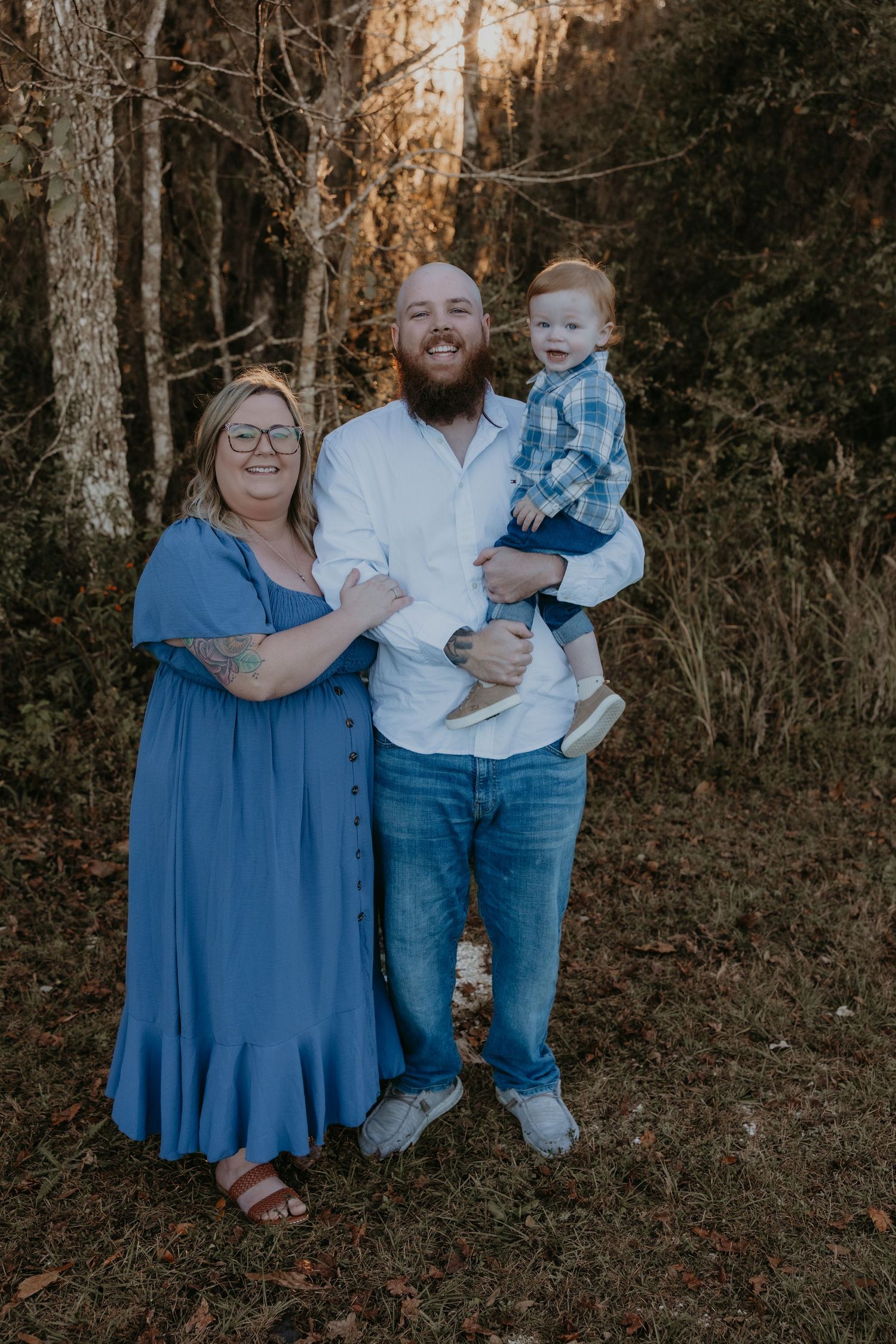 Family portrait of smiling couple with bearded man holding toddler boy, posing outdoors in blue outfits near autumn trees.