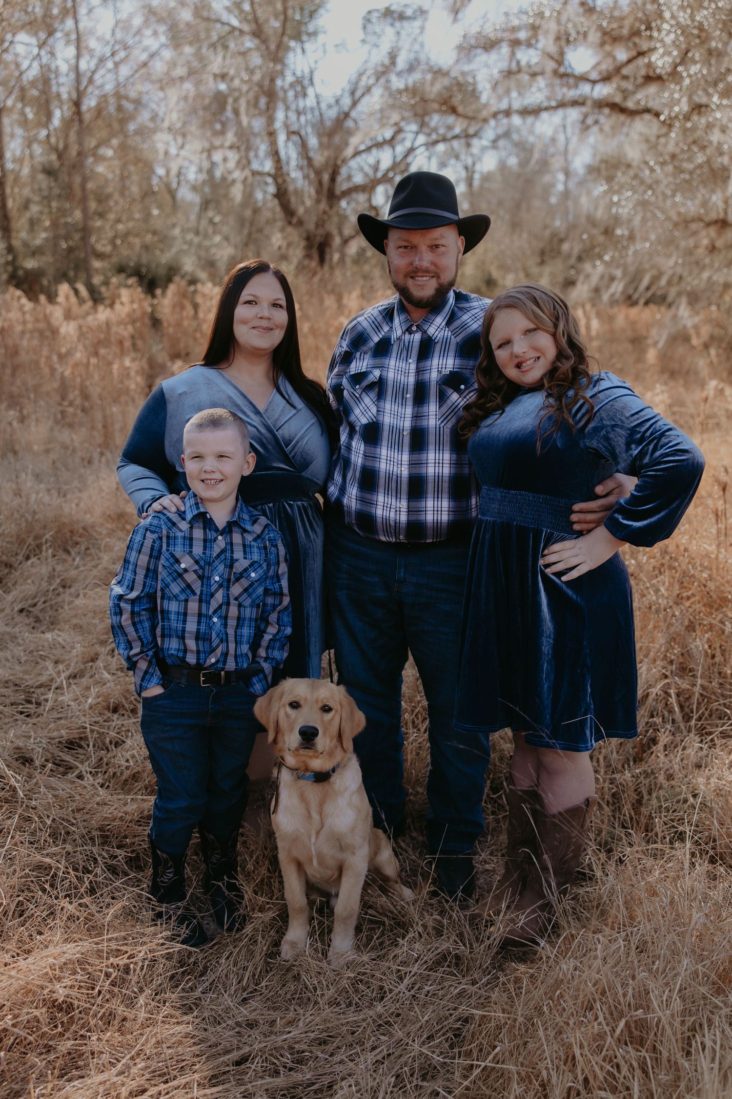 Family of four in coordinating blue outfits poses with a yellow Labrador in a rustic autumn field setting.