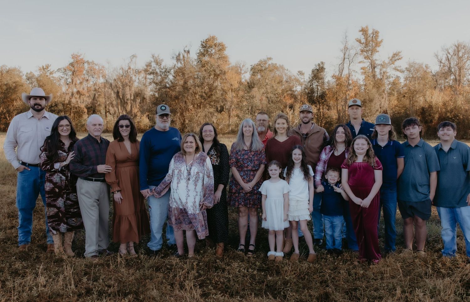 Large family group portrait outdoors in a field with autumn trees, dressed in coordinated outfits for a fall family photo session.
